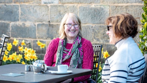 Two visitors enjoying a hot drink in the sunshine. The visitors are sitting at a table at the Clocktower Cafe (Wallington) and there are daffodils are in the background.
